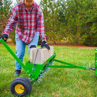 Dressed in a red plaid shirt, ripped jeans, and gloves, a person navigates a grassy area with the John Deere 40 Spike Aerator featuring a rigid steel weight tray. Steel tine stars aerate the earth as the trees sway gently in the background.