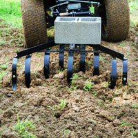 Close-up of a Brinly Parts Sleeve Hitch Cultivator | CC-560 attached to a small tractor, tilling and aerating garden soil. It features multiple curved metal tines and a concrete block for added weight, leaving loosened ground with patches of grass.