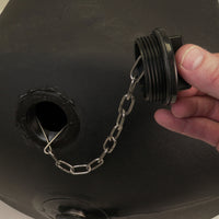 A hand holds the Brinly Parts 1019524 Tethered Plug, a black threaded plastic cap attached by a metal chain to a large black plastic container with an open threaded hole.