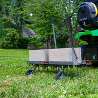 A lawn tractor with a Brinly Parts 40 Dethatcher in Hammered Gunmetal (DT2-40BH-S) and a black metal attachment, carrying cinder blocks, is parked on grassy ground. Trees and a building form a lush, serene backdrop.