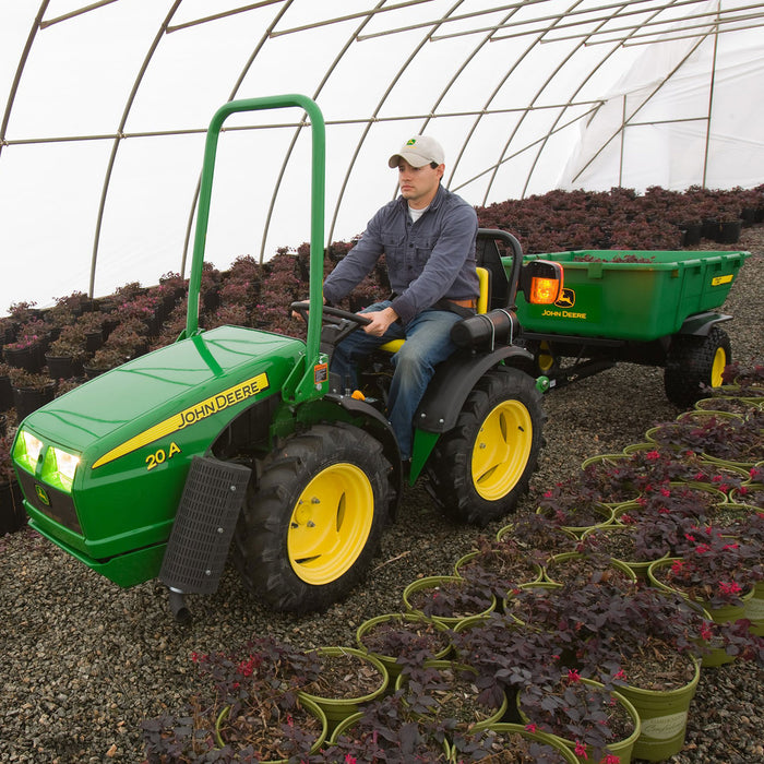 Inside a greenhouse, someone expertly maneuvers a green John Deere tractor with pneumatic tires among plant rows. The tractor pulls a John Deere 17 cu. Ft. Poly ATV Cart, filled with lush plants and lit by the natural light streaming through the curved, transparent structure.