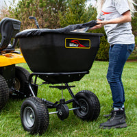 A person adjusts the cover on a rust-proof Brinly Parts 175 LB. Tow-Behind Deluxe Spreader (BS361BH-A) hitched to a yellow riding lawn mower on grass, with trees in the serene background.