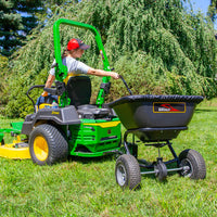 A person on a green ride-on lawn mower tows a rust-proof polyethylene hopper labeled Brinly Parts featuring the 125 LB. Tow-Behind Deluxe Spreader with Extended Handle & Cover (BS261BH-A) across a grassy lawn, wearing a red cap and gray shirt, with trees and bushes in the background.