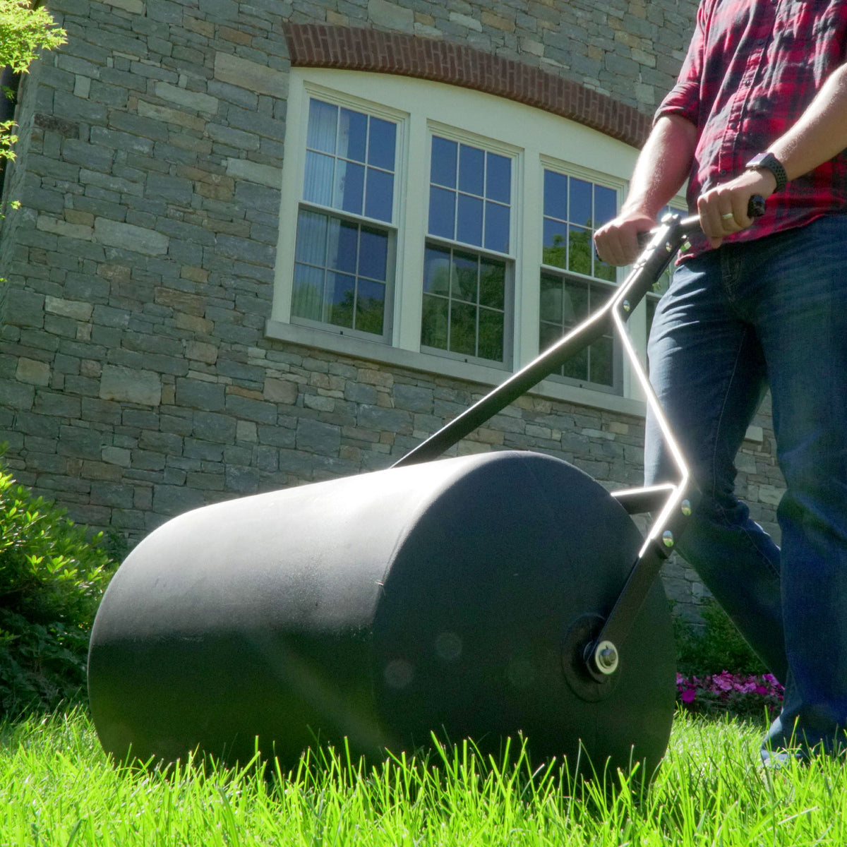 A person in jeans and a red shirt pushes a 28 Gallon Push or Tow Poly Lawn Roller (Brinly Parts PRC-242BH) across a green lawn in front of a stone house with white-trimmed windows. Bright sunlight highlights the lush grass and colorful flowers nearby.