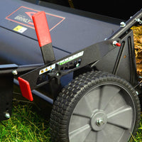 Close-up of the Brinly Parts 54″ Combo Lawn Sweeper with Dethatcher (LSDT2-54BH-G), showing its wheel and red-handled adjustment lever on green grass with dry leaves. The Brinly name appears on the machine.