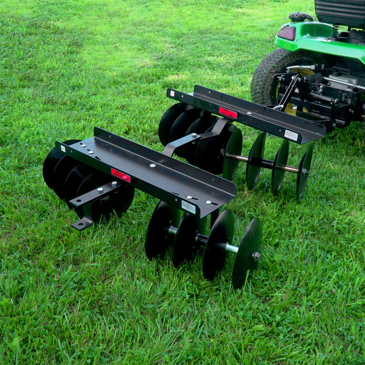 A Brinly Parts green tractor on a grassy lawn is equipped with the Sleeve Hitch Disc Harrow | DD-551BH, ideal for soil preparation with its black metal design and multiple circular blades, easily attached via a sleeve hitch for efficient cultivation.