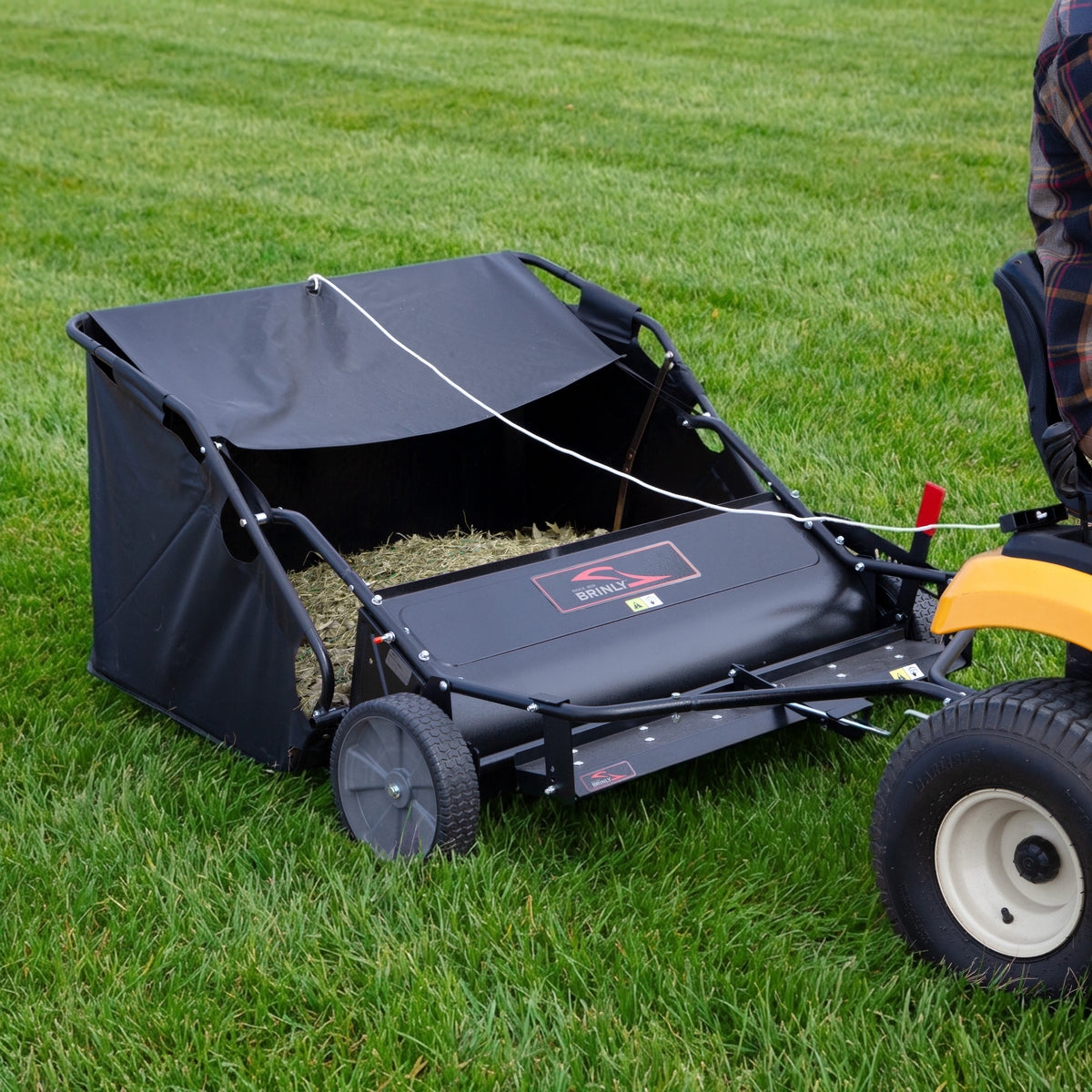 The Brinly Parts 54″ Combo Lawn Sweeper with Dethatcher (LSDT2-54BH-G) attaches to a riding mower, efficiently collecting lawn debris and grass clippings in its durable black wheeled catcher and fabric bag.