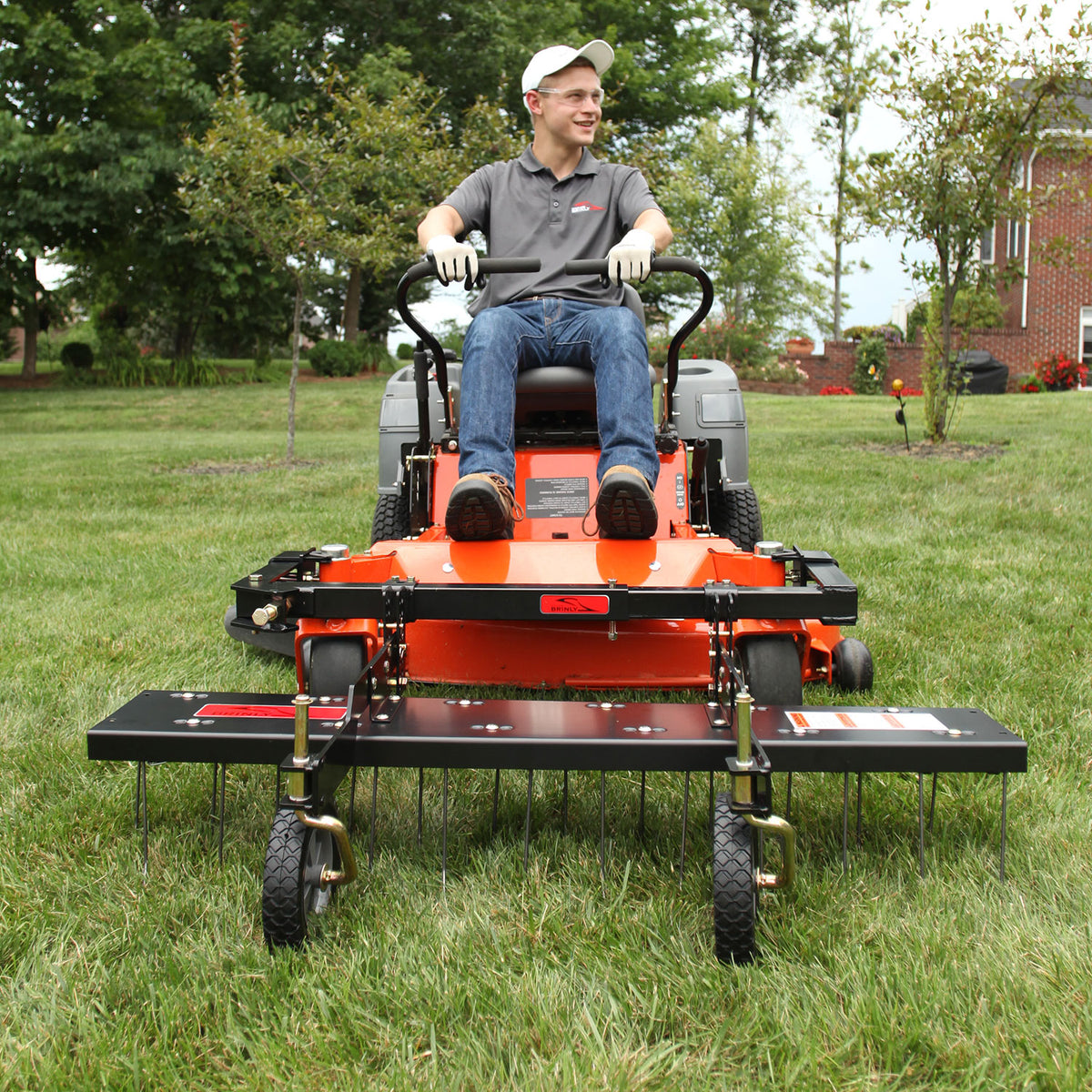 A person in a gray shirt, jeans, and cap sits on an orange mower in a grassy yard with trees and a brick building behind. The Brinly Parts 48” ZTR Dethatcher (DTZ-481BH), with extended wheels, highlights its lawn maintenance capabilities.