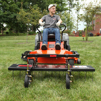 A person in a gray shirt, jeans, and cap sits on an orange mower in a grassy yard with trees and a brick building behind. The Brinly Parts 48” ZTR Dethatcher (DTZ-481BH), with extended wheels, highlights its lawn maintenance capabilities.