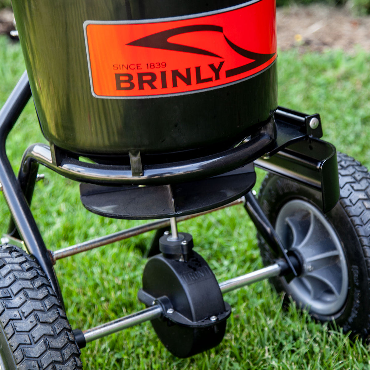 Close-up of a black and orange 50 LB. Brinly Parts P20-500BHDF push spreader with an optional deflector kit on green grass. Featuring two large wheels and a dispensing mechanism below, the label prominently displays the brand name and Since 1839.