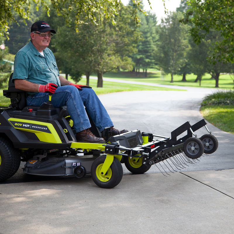 Sporting a cap and gloves, a person confidently sits on a Brinly Parts 48” ZTR Dethatcher | DTZ-481BH. On the paved driveway, this green and black machine stands ready for lawn maintenance, surrounded by lush grass and trees under a clear sky.