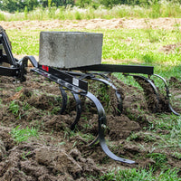 The Brinly Parts Sleeve Hitch Cultivator CC-560, with its curved metal tines and added weight from a concrete block, efficiently tills the soil on this sunny day, revealing aerated garden soil against lush green grass in the background.