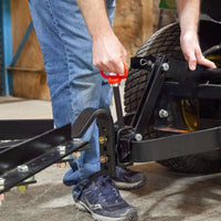 Wearing jeans and sneakers, a person uses a red-handled Brinly Parts Magnetic Hitch Pin - 5/8 Inch to secure a metal tow bar to a vehicle, preparing to attach tow-behind lawn equipment.