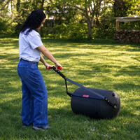 A person wearing a gray shirt and blue jeans pushes the Brinly Parts 28 Gallon Push or Tow Poly Lawn Roller (PRC-243BH) across a grassy yard, with trees and a woodpile in the background.