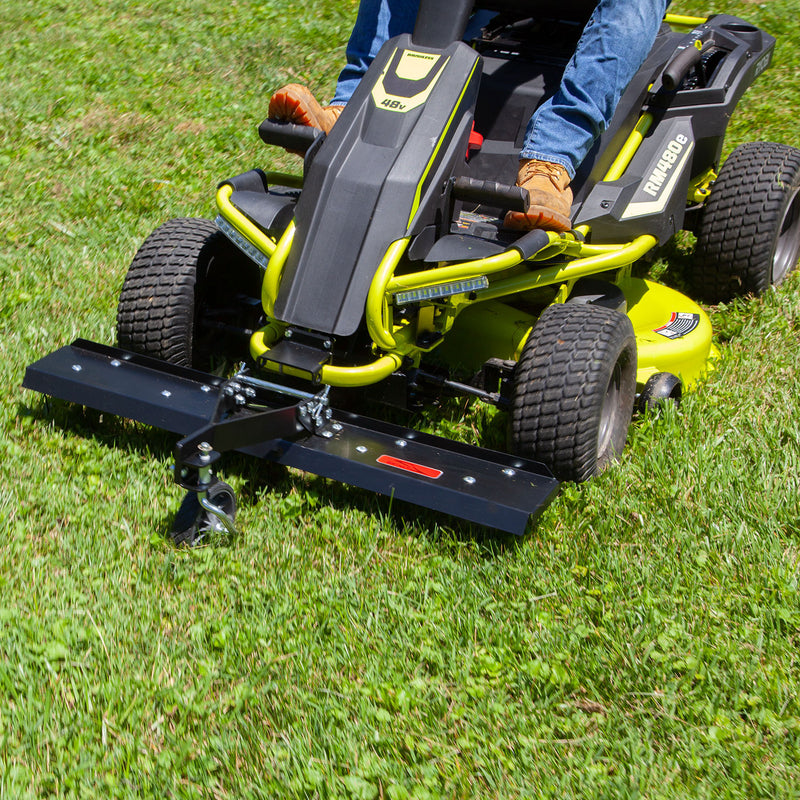 A person maneuvers a yellow RYOBI Electric Riding Mower, fitted with a Brinly Parts DT-38RY 38 in. Front Mount Dethatcher, across a grassy lawn. Its black frame and wide cutting deck leave precise tire treads for flawless lawn care.