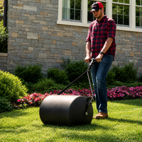 A person in a red plaid shirt and cap uses the Brinly Parts 28 Gallon Push or Tow Poly Lawn Roller (PRC-243BH) to roll over green grass in a landscaped yard with bushes and pink flowers, in front of a stone house.