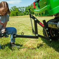 A person in jeans, boots, and a gray t-shirt kneels on grass, attaching the Brinly Parts Universal ATV/UTV One-Point Lift to the back of a green 4x4. Trees and fields stretch under a clear blue sky.