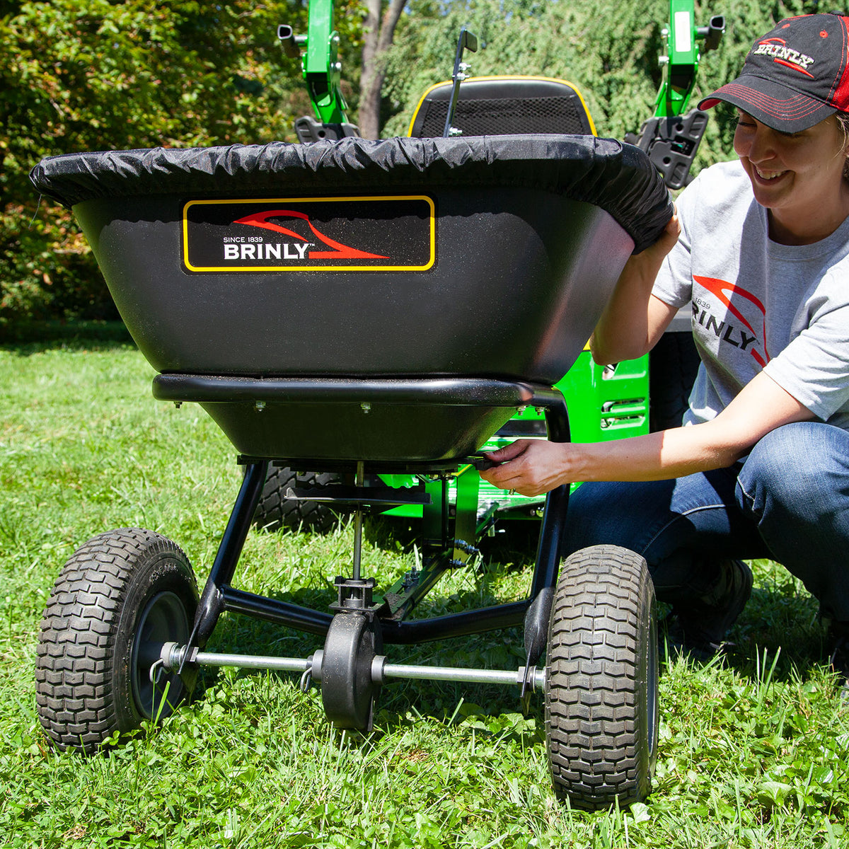 A person in a gray shirt and matching cap adjusts a black Brinly Parts 125 LB. Tow-Behind Deluxe Spreader (BS261BH-A) attached to a green tractor on grass, ready for efficient lawn fertilization.