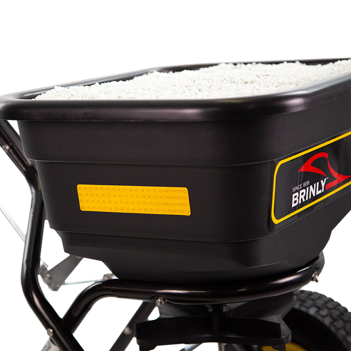 A close-up of Brinly Parts 70 lbs. Capacity Broadcast Ice Melt Spreader (PS10-70BH) shows a black spreader filled with white granules, featuring a yellow reflective strip and red and white logo on its side. It includes a wheeled cart and is designed for convenience in all conditions.