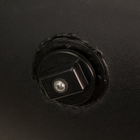 Close-up of the Brinly Parts 1019524 Tethered Plug, featuring a circular locking mechanism with a keyhole at the center and protruding square knob, set on a dark surface with a textured edge.